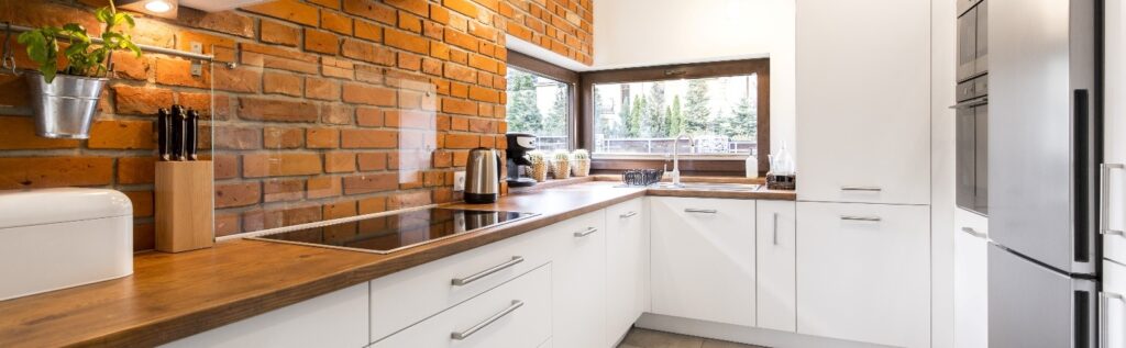 A kitchen with a brick wall, wooden counters, and white cabinetry.